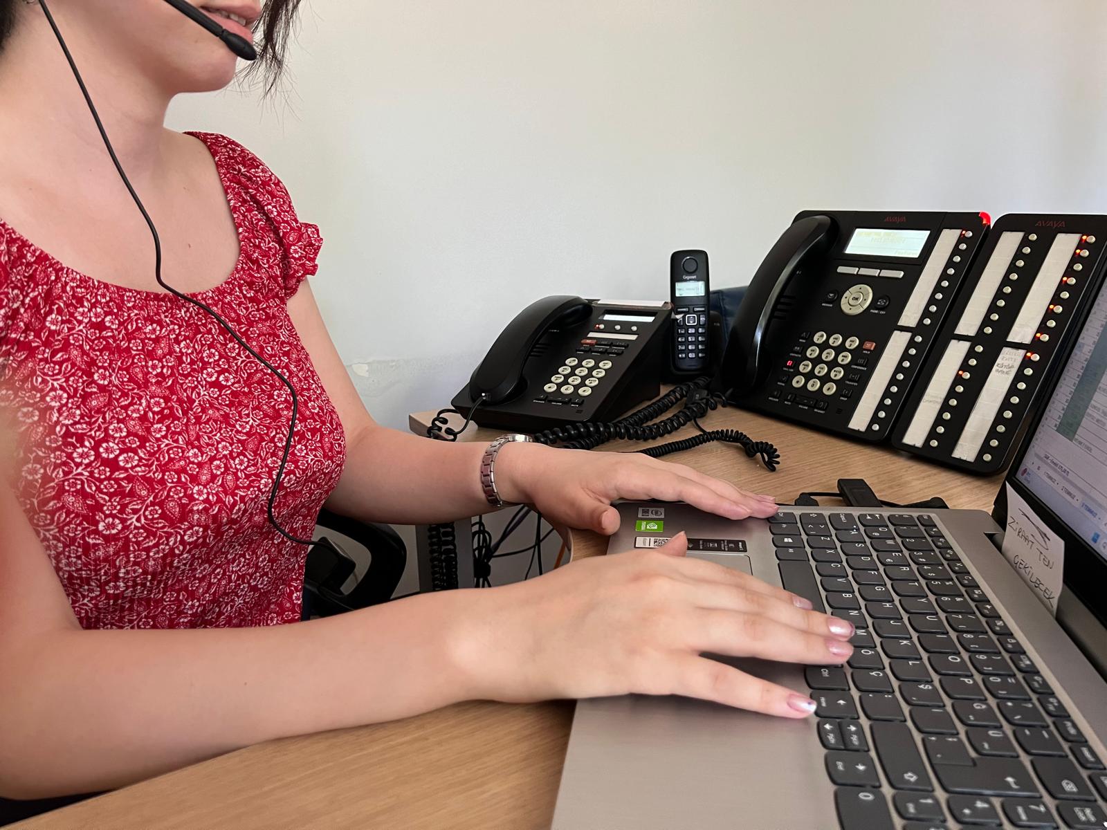 Fotoğrafta Rehabilitasyon Merkezi Santral sınıfında kahverengi masa üzerinde soldan sıralı olarak, bir sabit telefon, telsiz telefonu ve santral telefonu yer alıyor. Kablolu çağrı merkezi kulaklığı ve mikrofonu kullanan, yüzü bilgisayara dönük kırmızı tişörtlü genç bir kadının elleri bilgisayar klavyesine dokunuyor.