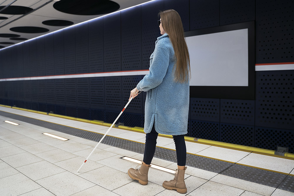 Fotoğrafta metro durağında bastonu ile yürüyen, kameraya arkası dönük mavi uzun mont, siyah tay ve bej renkli uzun konçlu botlar giyen uzun kumral saçları bulunan genç bir kadın yer alıyor.